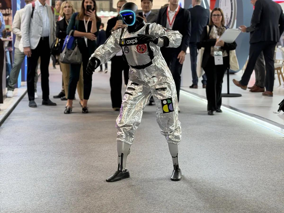 LéoBot, ein humanoider Roboter im silbernen Overall, in Tanzpose mitten in einem Messegang; hinter ihm bleiben Besucher stehen, schauen zu und filmen.