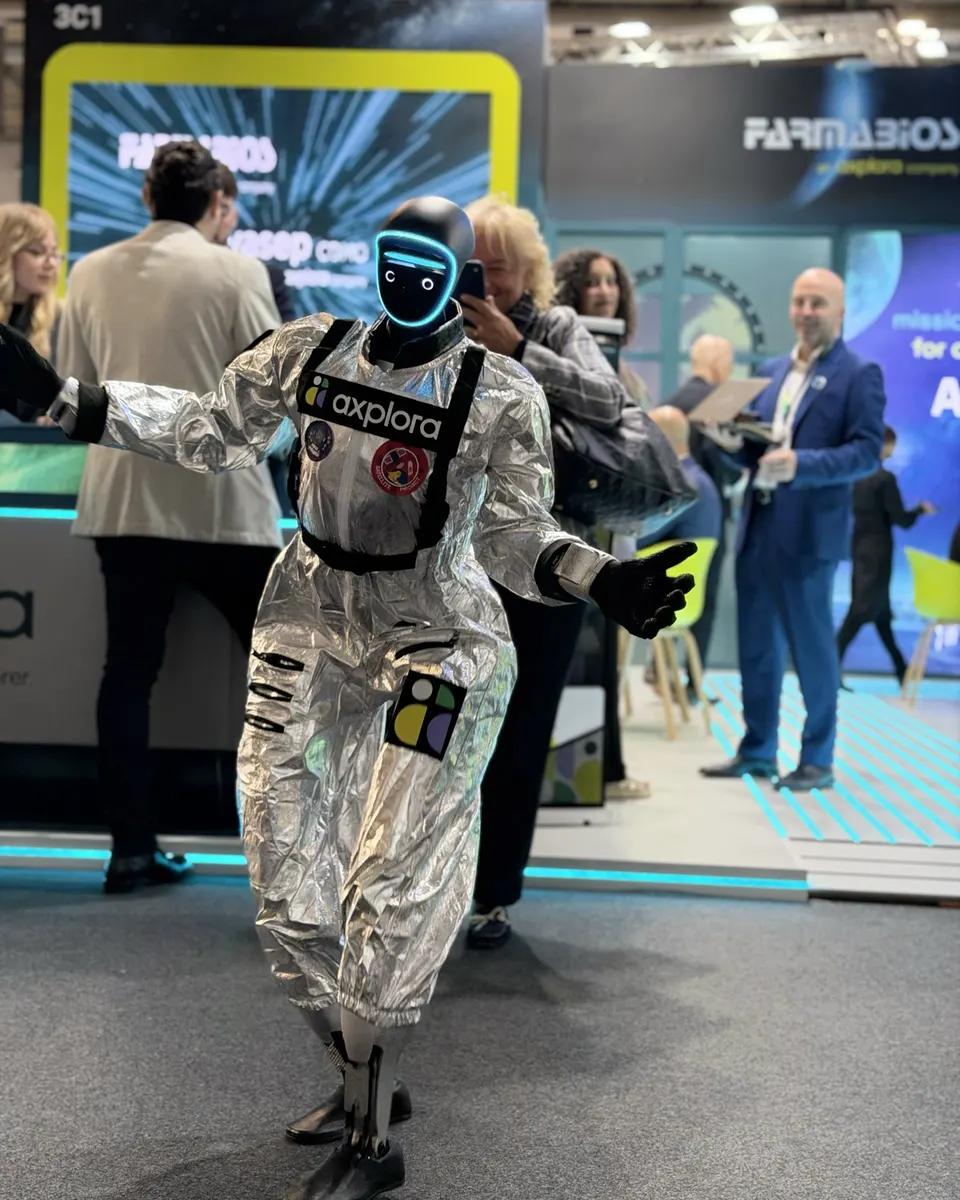 LéoBot tanzt mit ausgebreiteten Armen vor dem Tresen eines Messestands; hinter ihm fotografiert ihn eine lächelnde Besucherin mit dem Smartphone.